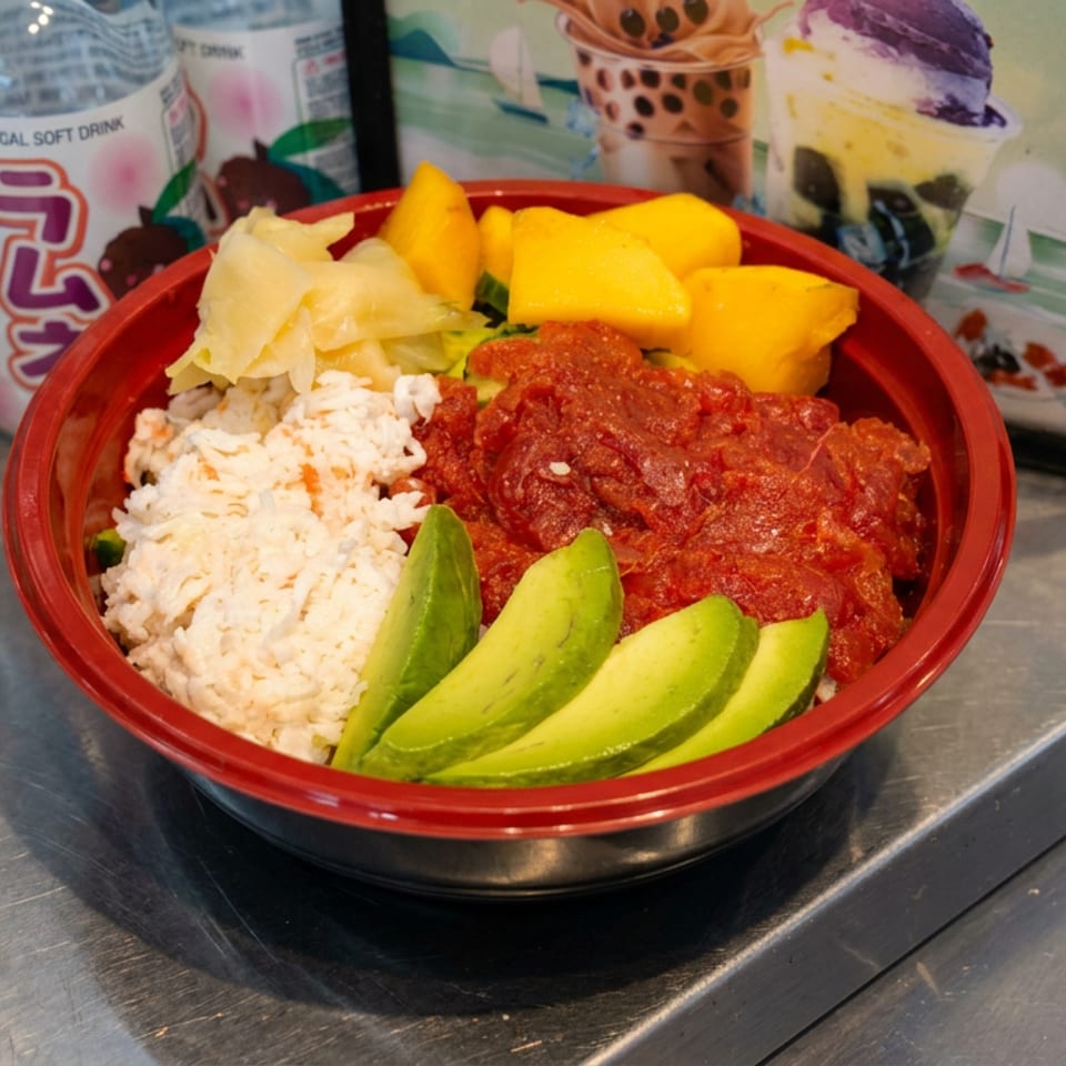 Bowl of white rice, spicy red chicken, avocado slices, cabbage, and mango on a restaurant counter beside bottled drinks.